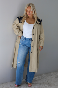 A woman wearing a tan trench coat with a navy collared neckline, functional button closures in the front, functional pockets on either side, with a tie waistline, paired with a white tank and light wash flared hem denim jeans, standing indoors against a white wall with with white marble flooring. 