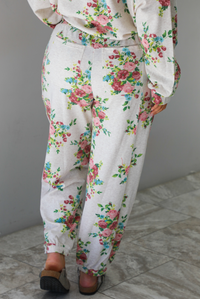 Woman wearing an oatmeal colored and multi-colored floral print two piece set featuring a collared long sleeved crop top and high waisted pants. The photo shows the back of the pants. The photo is taken indoors against a white wall with white marble flooring. 
