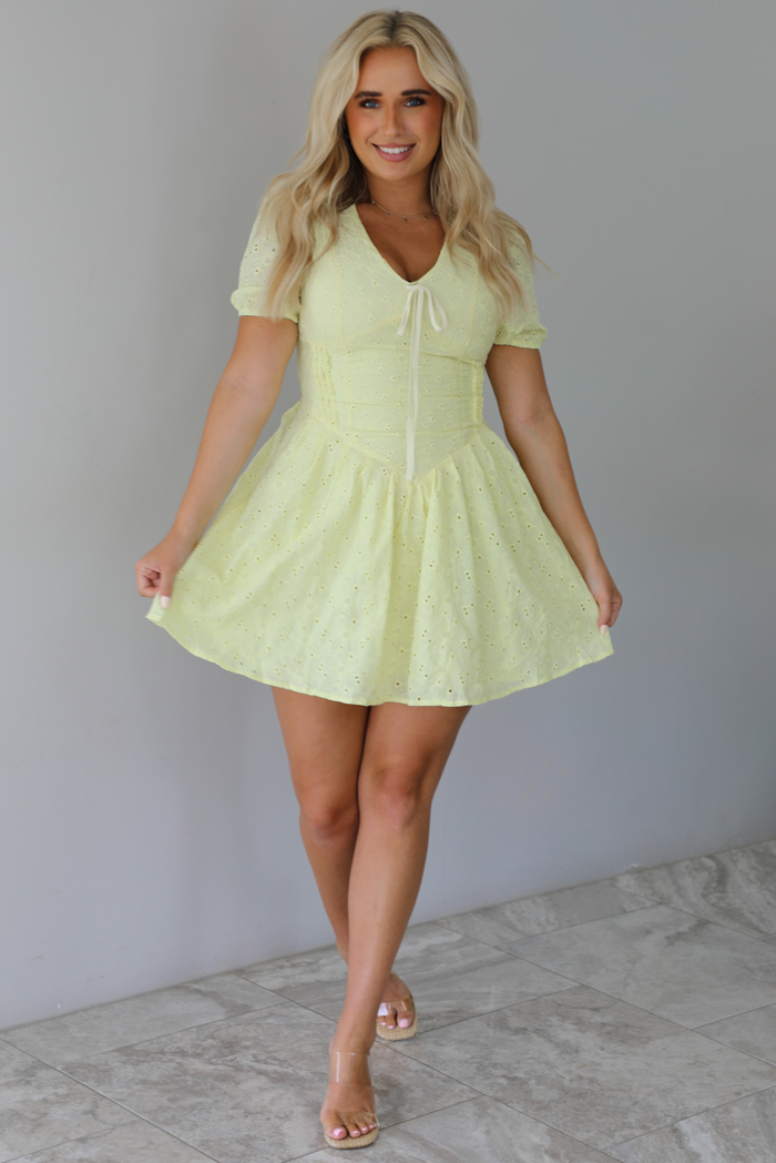 Woman wearing a light yellow mini dress with a floral eyelet design throughout, v-neckline, short sleeves, fitted drop waist bodice, and a flowy mini length skirt indoors against a white wall with white marble flooring. 