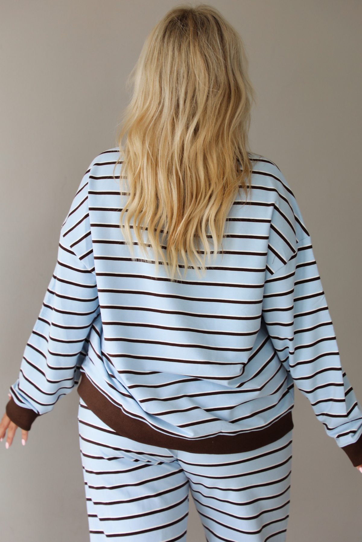 A woman shown from behind wearing a blue matching two piece set with dark brown and white striped details, featuring an oversized long sleeved top and high waisted loose fitting, flared leg pants, indoors against a white wall. The model has the top untucked, showing the full length of the back of the top, wth the brown hem detail on both the bottom hem of the top and the wrists. 