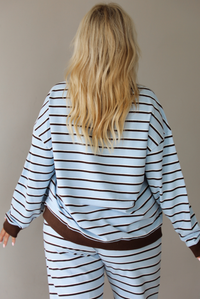 A woman shown from behind wearing a blue matching two piece set with dark brown and white striped details, featuring an oversized long sleeved top and high waisted loose fitting, flared leg pants, indoors against a white wall. The model has the top untucked, showing the full length of the back of the top, wth the brown hem detail on both the bottom hem of the top and the wrists. 