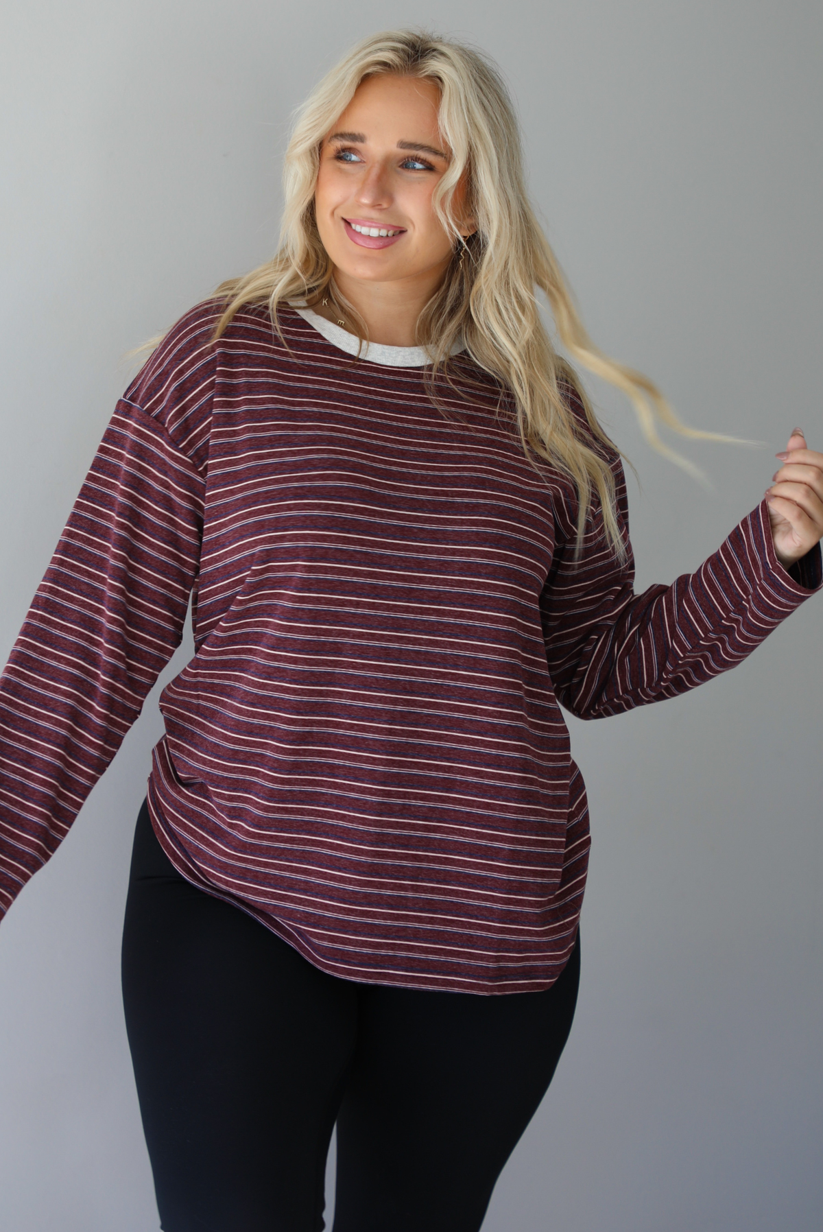 A woman wearing a burgundy striped long sleeved top paired with black leggings, standing indoors against a white wall. 
