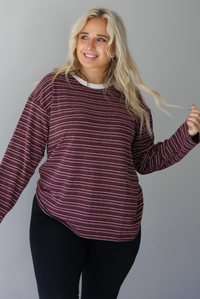 A woman wearing a burgundy striped long sleeved top paired with black leggings, standing indoors against a white wall. 