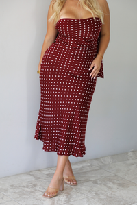A woman wearing a strapless maroon midi dress with a white polka dot pattern with a matching scarf, standing indoors against a white wall with white marble flooring. 