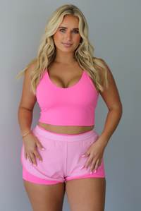 A woman modeling a bright pink two-piece activewear set consisting of a scoop-neck crop top and matching athletic shorts with a darker, hot pink trim, standing indoors against a white wall.