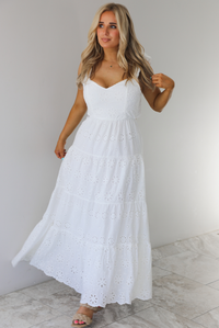 Woman wearing a white maxi length dress that has an eyelet floral design throughout and has a fitted bodice. The photo is taken indoors against a white wall with white marble flooring. 