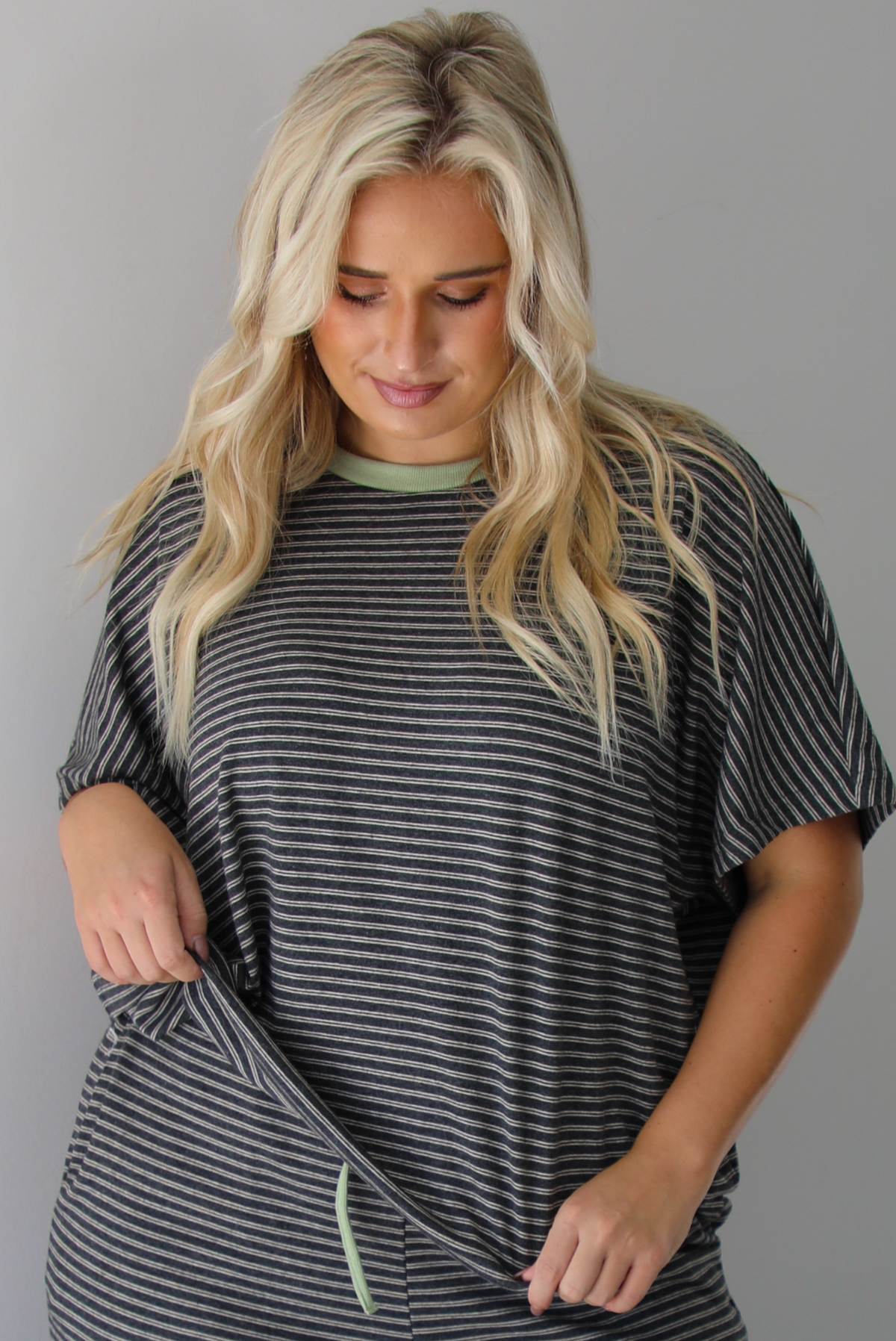 A woman wearing a charcoal and white striped two piece matching set with green trim detailing at the neckline and waistline, featuring an oversized short sleeved top and high waisted pants, standing indoors against a white wall. The photo shows a closeup of the top. 