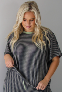 A woman wearing a charcoal and white striped two piece matching set with green trim detailing at the neckline and waistline, featuring an oversized short sleeved top and high waisted pants, standing indoors against a white wall. The photo shows a closeup of the top. 