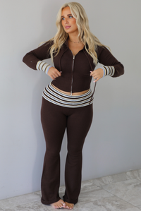 A woman modeling a dark brown ribbed zip-up hoodie and matching flared pants set with white striped accents on the cuffs and waistband, standing indoors against a white wall with white marble flooring. 