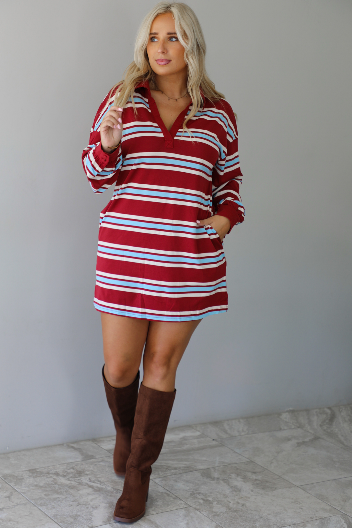 A woman wearing a red burgundy mini dress with a white and blue striped pattern throughout, collared neckline, long sleeves, and functional pockets on ether side, standing indoors against a white wall with white marble flooring. 