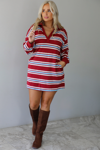 A woman wearing a red burgundy mini dress with a white and blue striped pattern throughout, collared neckline, long sleeves, and functional pockets on ether side, standing indoors against a white wall with white marble flooring. 
