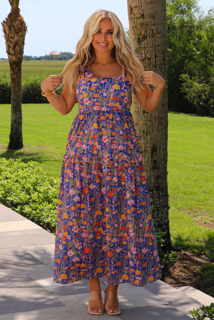 A woman wearing a sleeveless tiered navy blue maxi dress with a colorful floral print with a fitted bodice and flowy skirt, standing outdoors on a paved walkway next to a palm tree.
