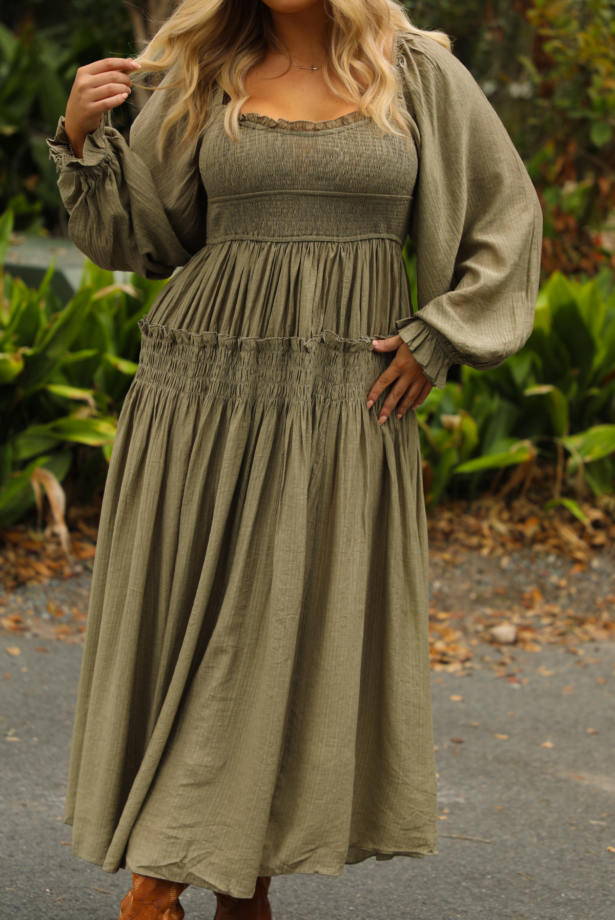 Close up of a woman model wearing an olive colored maxi length dress that has a smocked bodice, long sleeves that cinch with elastic at the wrists, and a flowy skirt. The photo is taken outdoors with greenery in the back.