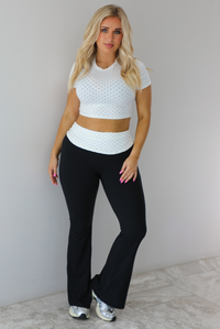 Woman wearing a two piece set featuring a white cropped top with a black polka dot print pattern and black pants that are high waisted with a white and black polka dot print folded over detail. The photo is taken indoors against a white wall with white marble flooring.