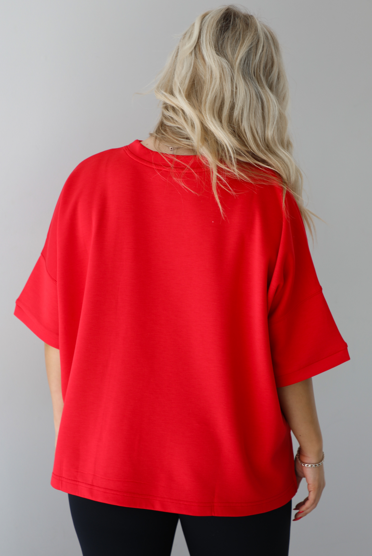 Woman wearing an oversized red t-shirt with black, fitted lettings. The photo shows a closeup of the back of the top. The photo is taken indoors against a white wall.