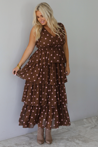 A woman wearing a brown midi length ruffled dress with a pink polka dot print pattern throughout, standing indoors against a white wall with white marble flooring. 
