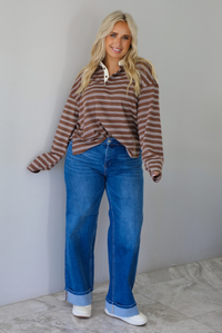 A woman wearing a brown and cream striped collared quarter-button long sleeved top, front tucked into dark wash denim wide leg jeans that have a cuffed hem, paired with cream sneakers, standing indoors against a white wall with white marble flooring. 