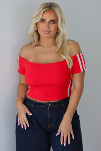 A blonde woman modeling a bright red off-the-shoulder short-sleeve top with white stripes on the sleeves, paired with dark wide-leg denim jeans, standing indoors against a white wall.