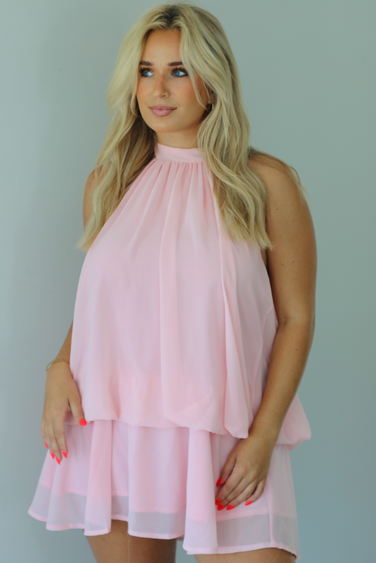 Woman wearing a pink dress that has a halter tie neckline, a cinched detail at the hips, and a flowy design indoors against a white wall.