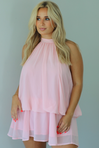 Woman wearing a pink dress that has a halter tie neckline, a cinched detail at the hips, and a flowy design indoors against a white wall.