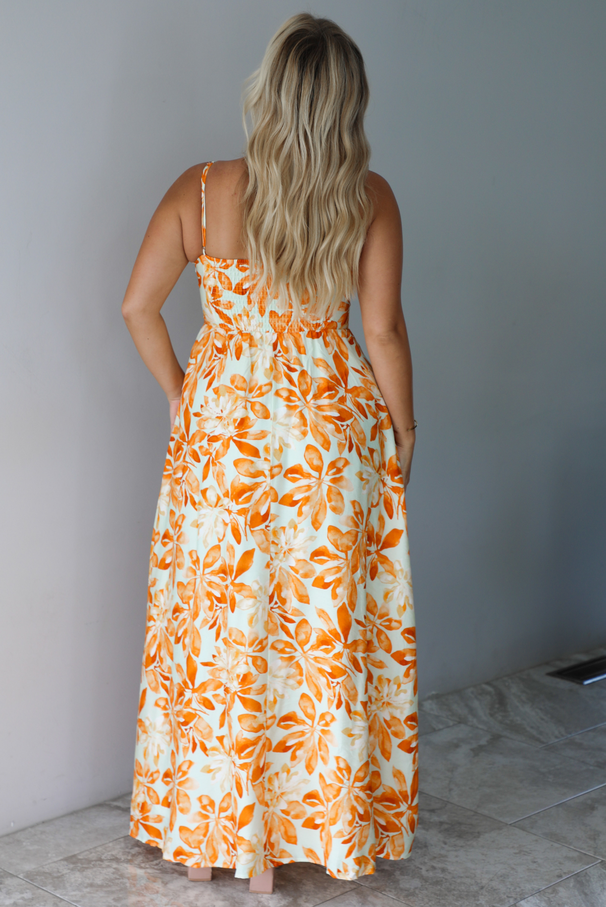 A woman shown from behind modeling a spaghetti strapped, floor-length maxi dress with a white base and a vibrant marigold orange floral pattern, standing indoors against a white wall with white marble flooring. The photo shows the back of the dress, focusing on the smocked detail at the back and adjustable straps. 