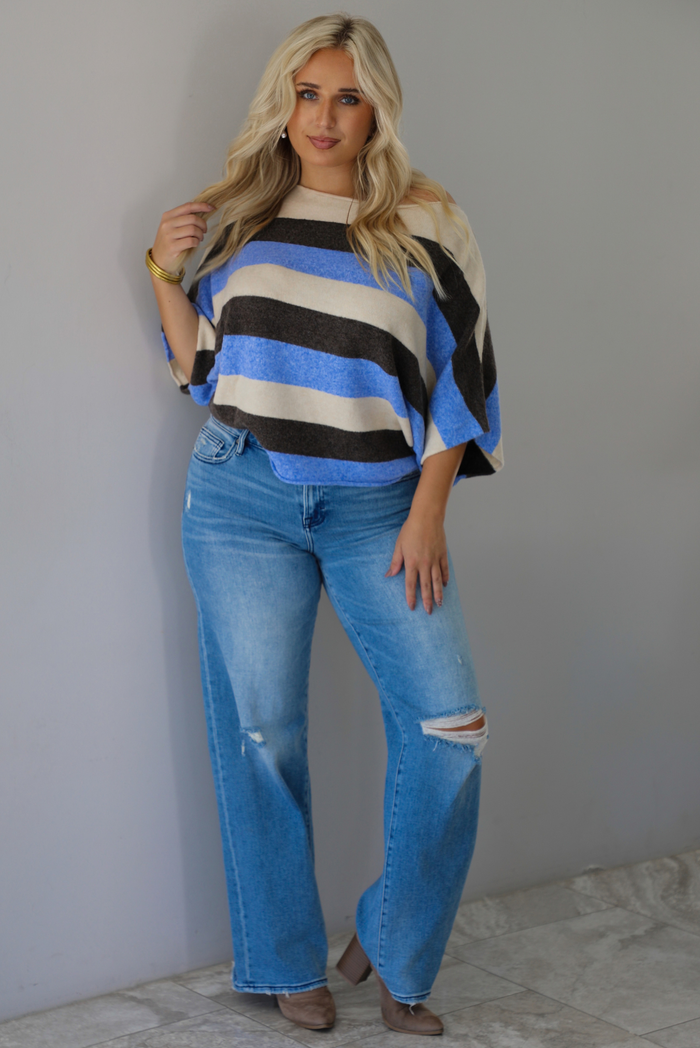 A woman wearing a cream, brown,, and blue striped knit top that has a boat-neck design, and quarter length flowy sleeves, paired with a pair of medium wash denim jeans, standing indoors against a white wall with white marble flooring. 
