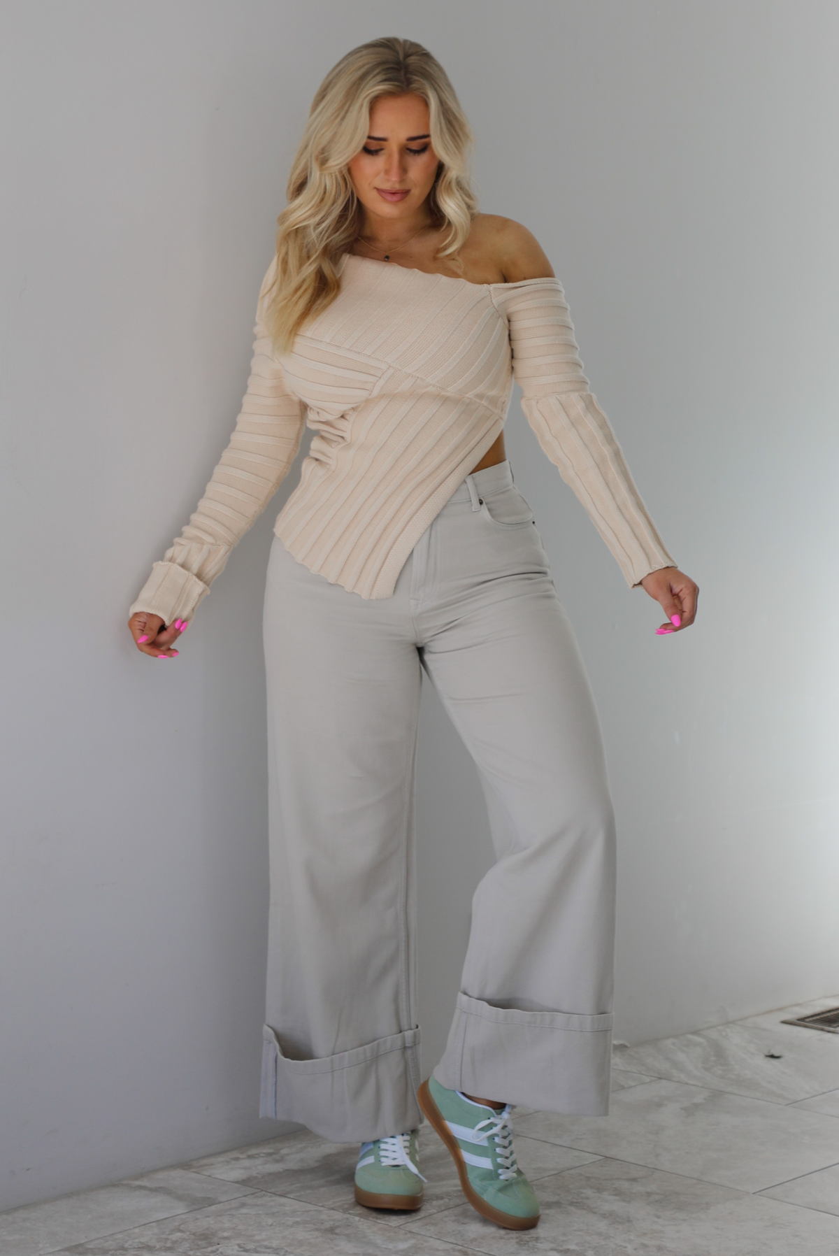 Woman wearing a cream colored ribbed textured knit top that has an asymmetrical hem with long sleeves in which one side is worn off the shoulder. The sweater is paired with sand colored high waisted, cuffed hem denim. The photo is taken indoors against a white wall with white marble flooring. 