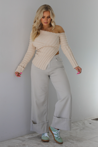 Woman wearing a cream colored ribbed textured knit top that has an asymmetrical hem with long sleeves in which one side is worn off the shoulder. The sweater is paired with sand colored high waisted, cuffed hem denim. The photo is taken indoors against a white wall with white marble flooring. 