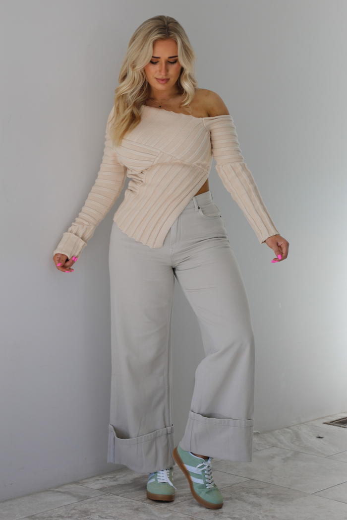 Woman wearing a cream colored ribbed textured knit top that has an asymmetrical hem with long sleeves in which one side is worn off the shoulder. The sweater is paired with sand colored high waisted, cuffed hem denim. The photo is taken indoors against a white wall with white marble flooring. 