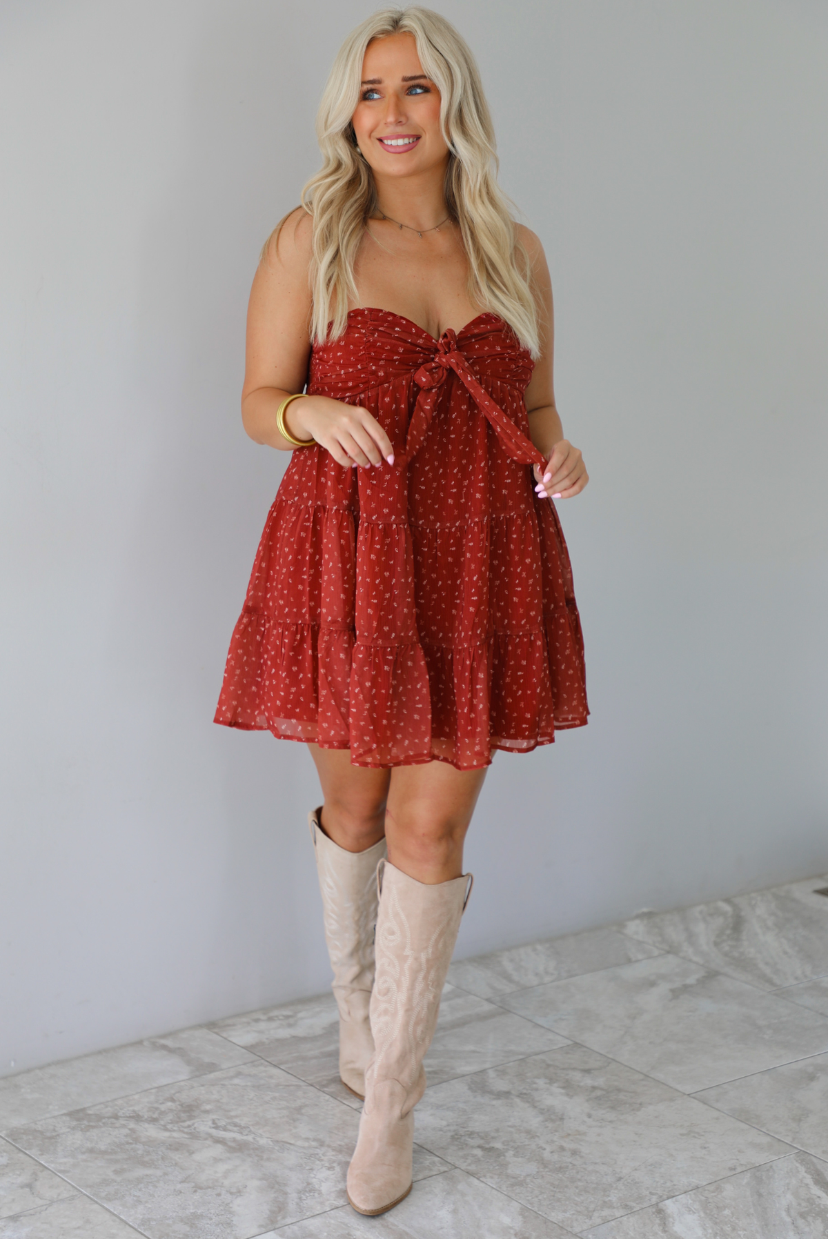 A woman wearing a rust mini length dress with a cream floral and leaf tiny print design throughout, has a strapless design with a tie detail in the center of the bust, with a baby-doll dress cut that's fitted at the bust with a flowy skirt, standing indoors against a white wall with white marble flooring. 