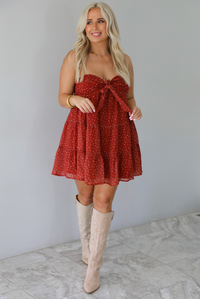 A woman wearing a rust mini length dress with a cream floral and leaf tiny print design throughout, has a strapless design with a tie detail in the center of the bust, with a baby-doll dress cut that's fitted at the bust with a flowy skirt, standing indoors against a white wall with white marble flooring. 