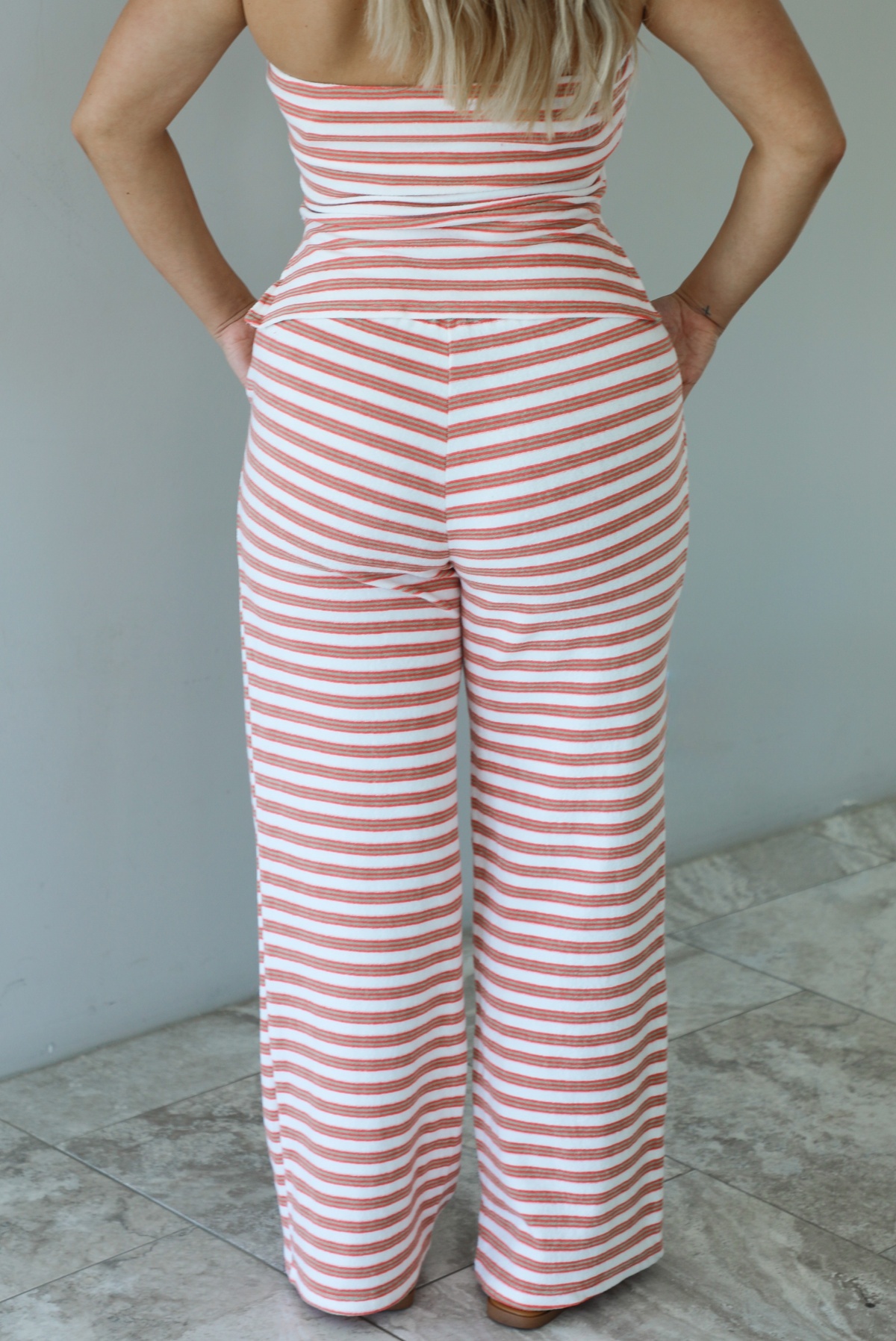 Woman wearing an orange, white, and green striped pattern set that has a strapless tube top and high waisted pants indoors against a white wall with white marble flooring and shows a closeup of the back of the pants, which have an elastic and drawstring waistline with functional pockets on either side. 