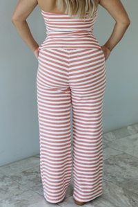 Woman wearing an orange, white, and green striped pattern set that has a strapless tube top and high waisted pants indoors against a white wall with white marble flooring and shows a closeup of the back of the pants, which have an elastic and drawstring waistline with functional pockets on either side. 