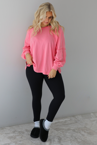 Woman wearing a pink oversized long sleeved top that has a high neckline, exposed seam details, split hem details on either side, and is lightweight. The top is modeled with a pair of fitted black leggings with black uggs and socks. The photo is modeled indoors against a white wall with white marbled flooring. 