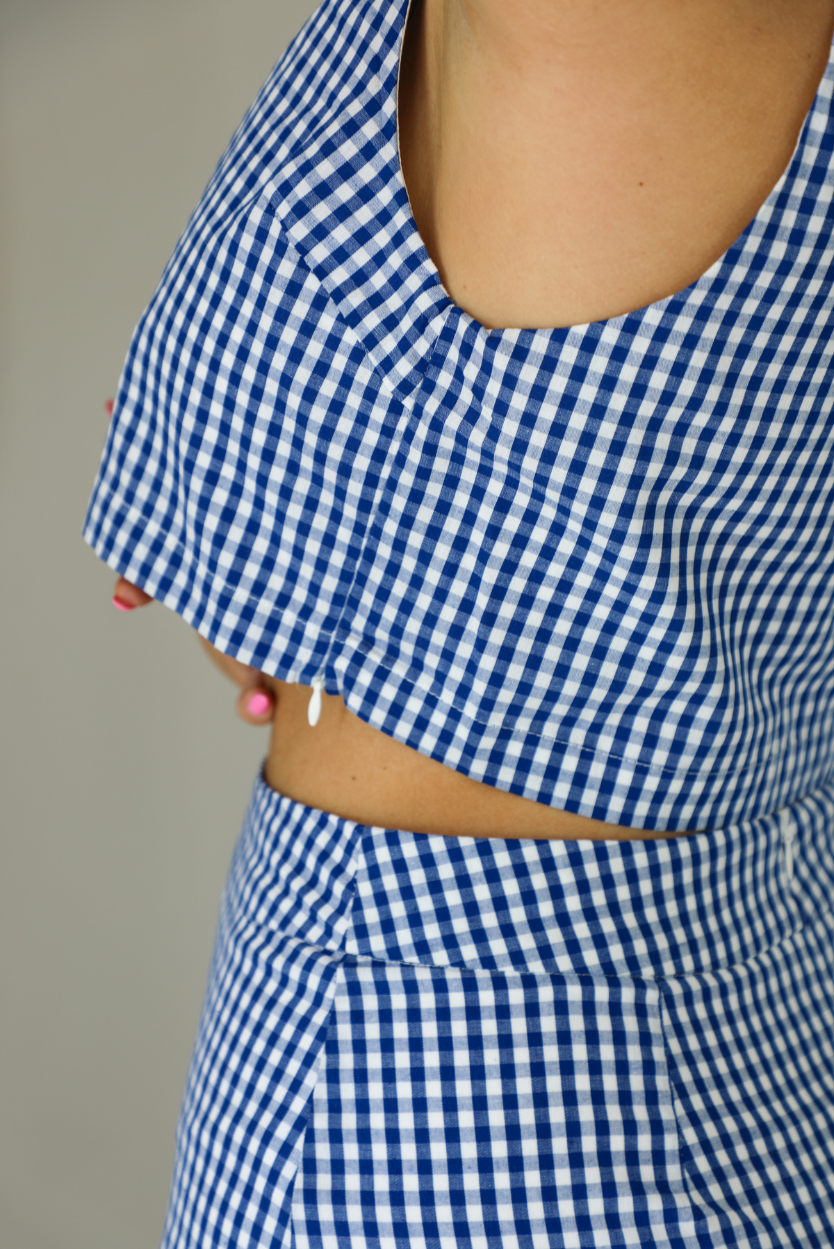 Woman wearing a blue and white gingham print patterned set featuring a cropped top and high waisted shorts that have a white peek-a-boo shorts liner underneath. The photo shows a closeup of the zipper closure on the left side of the top. The photo is taken indoors against a white wall.