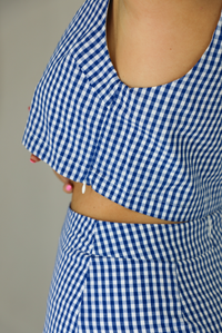 Woman wearing a blue and white gingham print patterned set featuring a cropped top and high waisted shorts that have a white peek-a-boo shorts liner underneath. The photo shows a closeup of the zipper closure on the left side of the top. The photo is taken indoors against a white wall.