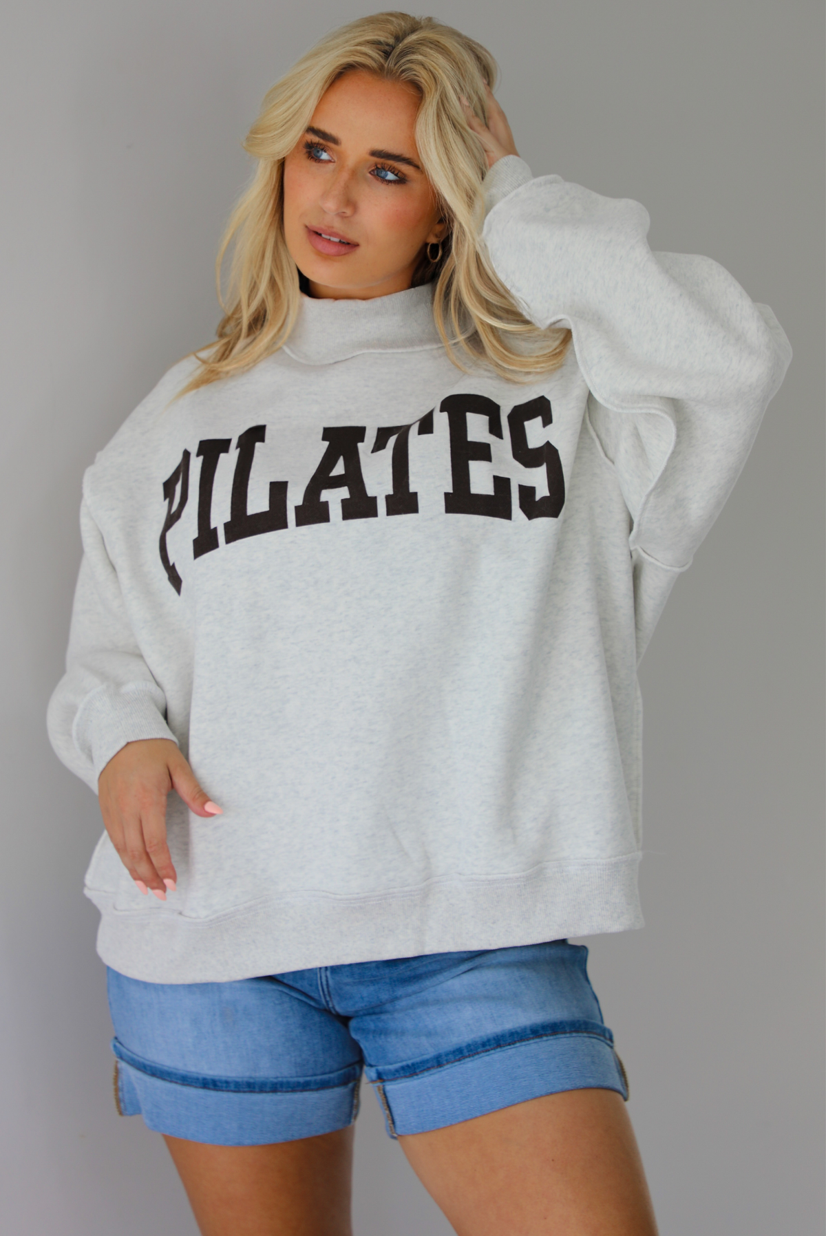 Woman wearing a heather grey oversized sweatshirt with the word 'PILATES' written in collegiate font on the front with medium wash denim shorts indoors against a white wall.