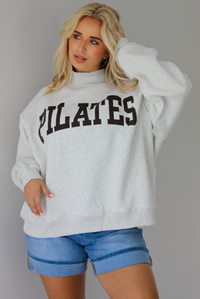 Woman wearing a heather grey oversized sweatshirt with the word 'PILATES' written in collegiate font on the front with medium wash denim shorts indoors against a white wall.