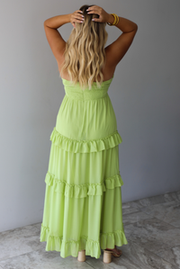 A woman shown from behind modeling a lime green halter-neck maxi dress with tiered ruffle details down the skirt, standing indoors against a white wall.