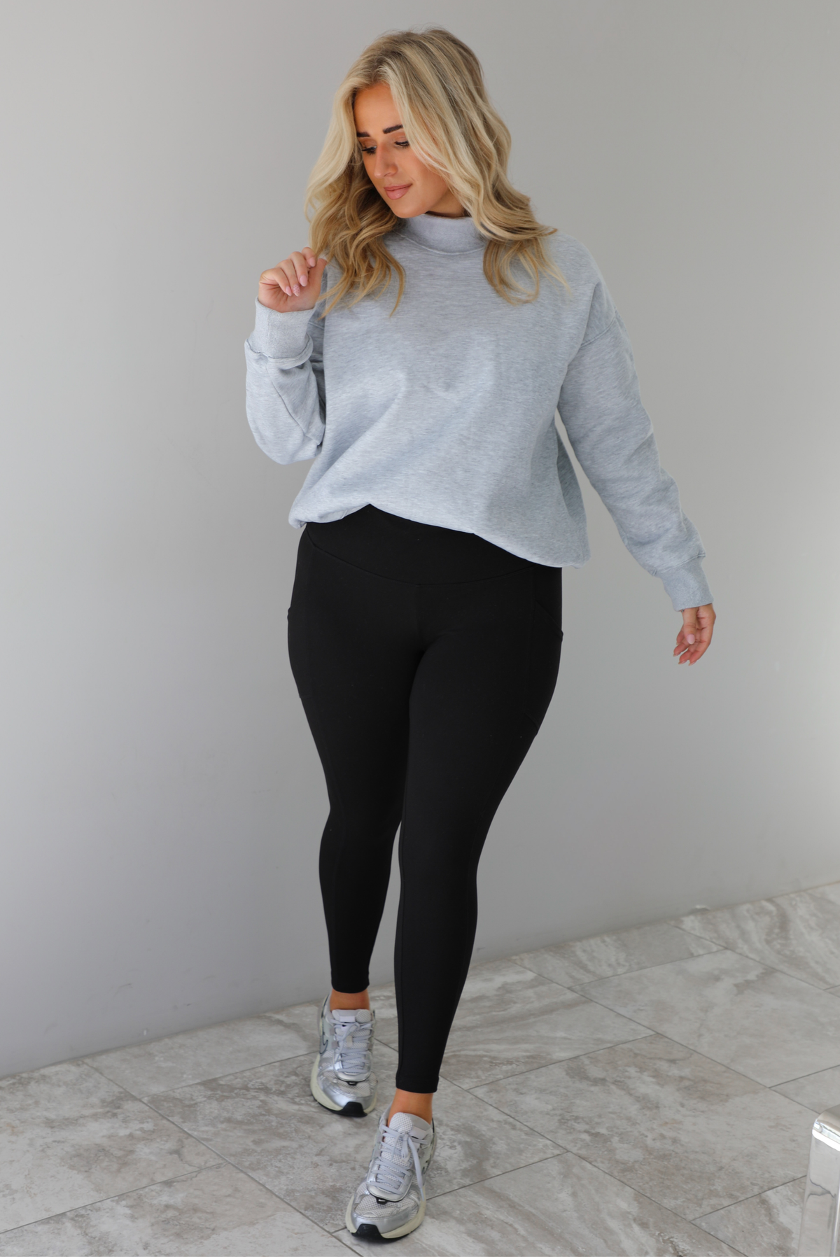 Woman wearing a grey turtleneck sweater tucked into high waisted black leggings. The outfit is paired with silver sneakers. The photo is taken indoors against a white wall with white marble flooring.