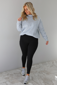 Woman wearing a grey turtleneck sweater tucked into high waisted black leggings. The outfit is paired with silver sneakers. The photo is taken indoors against a white wall with white marble flooring.