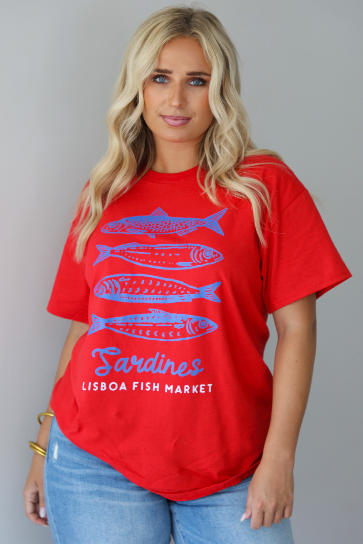A woman wearing a red graphic t-shirt with blue sardine illustrations and the text 'Sardines LISBON FISH MARKET', paired with blue jeans, standing indoors against a white wall.