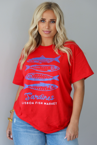A woman wearing a red graphic t-shirt with blue sardine illustrations and the text 'Sardines LISBON FISH MARKET', paired with blue jeans, standing indoors against a white wall.