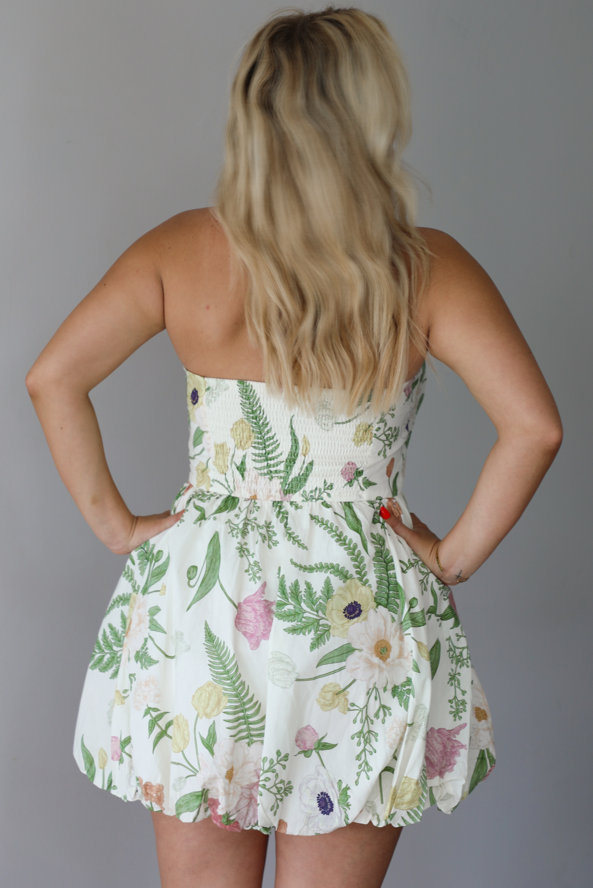 Woman wearing a cream strapless mini dress that has a multi-colored floral pattern throughout indoors against a white wall. The photo shows the back of the dress. 
