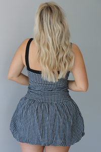 Woman wearing a black and white gingham print mini dress with a fitted, drop waist design, and a puffed hem skirt indoors against a white wall. The photo shows the back of the dress. 