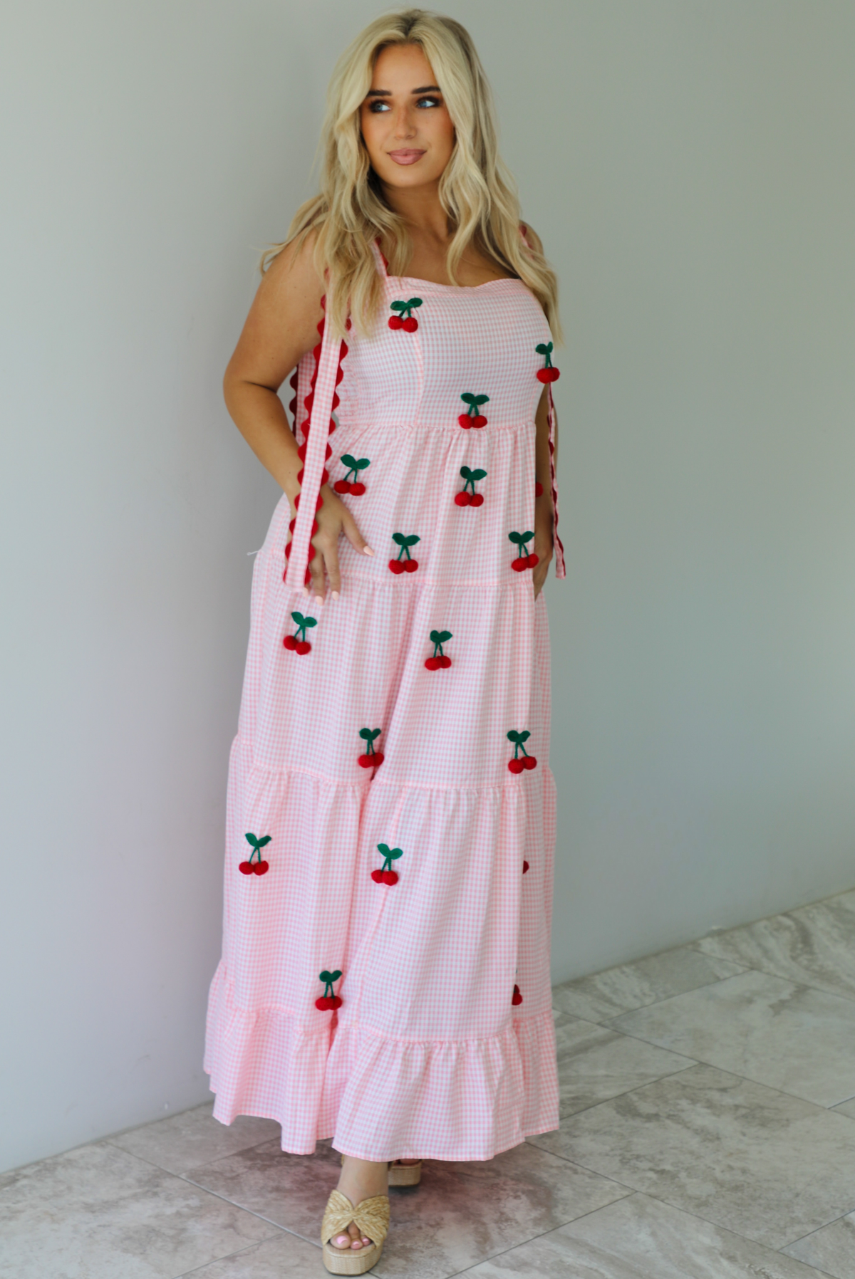 Woman wearing a pink and white gingham  maxi dress with thick straps, a fitted bodice, flowy skirt, and crocheted cherry details on the front indoors against a white wall with white marble flooring. 