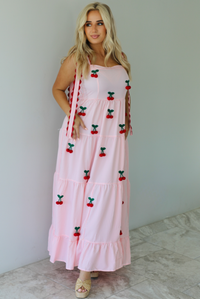 Woman wearing a pink and white gingham  maxi dress with thick straps, a fitted bodice, flowy skirt, and crocheted cherry details on the front indoors against a white wall with white marble flooring. 
