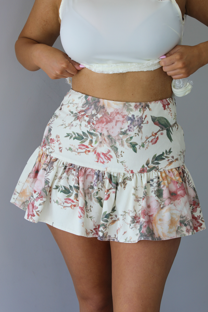 Woman wearing a cream and multi-colored floral skirt that has a high waistline, and a shorter length. The photo is taken indoors against a white wall.