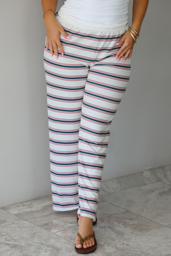 A person wearing wide-leg pants with horizontal green and red on a cream-white base, paired with a white top and brown sandals, standing indoors against a white wall with white marble flooring. 