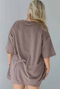 Woman wearing a two piece taupe set featuring an oversized t-shirt and fitted biker shorts. The photo shows the back of the oversized top. The photo is taken indoors in front of a white wall.